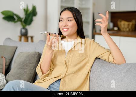 Frau, die die Spracherkennungs-App auf dem Mobiltelefon nutzt und im Mikrofon des Smartphones spricht. Charmante junge Frau, die zu Hause auf dem Sofa sitzt, Sprachnachrichten aufnimmt und sendet Stockfoto