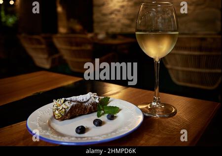 Sizilianische Cannoli Rollen mit Blaubeeren auf einer weißen Keramikplatte und ein Glas nebelverhangten Weißweins auf einem Restauranttisch mit verschwommenem Hintergrund. Stockfoto