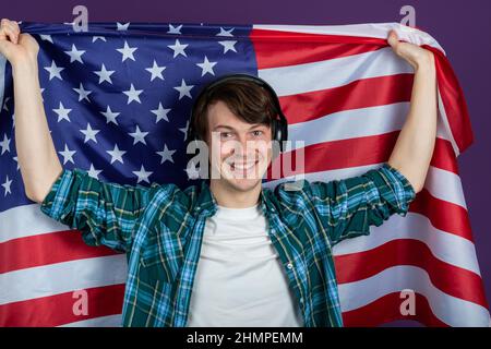 Porträt eines glücklichen Studenten im grünen Hemd, der Kopfhörer mit amerikanischer Flagge auf purpurem Hintergrund trägt und lächelt und die Kamera anschaut Stockfoto