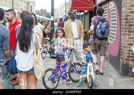 London, Großbritannien - 17. Juli 2020, Kinder, Geschwister auf Fahrrädern, hielten an, um Straßenmusikern auf dem Columbia's Sunday Flower Market zuzuhören Stockfoto