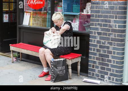 London, Großbritannien - am 17. Juli 2022 Sucht Ein modisch gekleidetes blondes Mädchen ein Telefon in ihrer Tasche und sitzt auf einer Bank in der Nähe des Geschäfts. Stockfoto