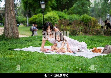 Charmante junge Mutter, die mit einem Kleinkind spielt und auf einer Decke im Park sitzt Stockfoto