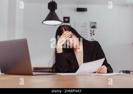 Geballte Frau in einer Brille, die das Dokument mit einem Blatt Papier liest, durchdachte Geschäftsfrau, freie Mitarbeiterin, die mit der Korrespondenz arbeitet und die Finanzlage überprüft Stockfoto