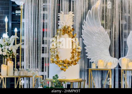 Blick aus der unteren Ecke auf den süßen Taufkuchen mit goldenen Ornamenten, Kerzen und Engelsflügeln im Restaurant Stockfoto