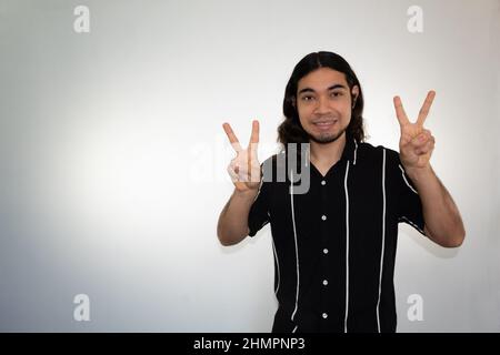 Porträt eines nicht-binären hispanischen Mannes, der das Friedenszeichen auf weißem Hintergrund macht Stockfoto