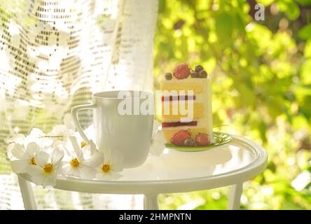 Schöne Porzellan-Kaffeetasse und Stück Kuchen auf Glas-Tisch mit weißen Blumen im Sommergarten im Sonnenlicht dekoriert. Speicherplatz kopieren. Sommergetränke-Konzept. Stockfoto