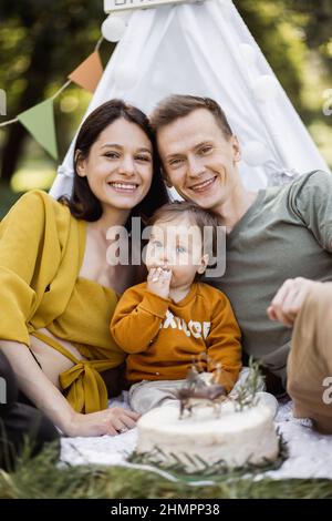 Porträt lächelnder junger Eltern, die ihren kleinen Sohn umarmen, der leckeren Geburtstagskuchen isst. Glückliche Familie von drei mit Geburtstagsfeier an der frischen Luft. Stockfoto