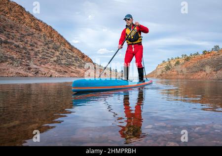 Senior männlichen Paddler in Trockentauchanzug ist Stand-up Paddeln auf See in Colorado, Winterlandschaft mit etwas Eis genießen. Stockfoto