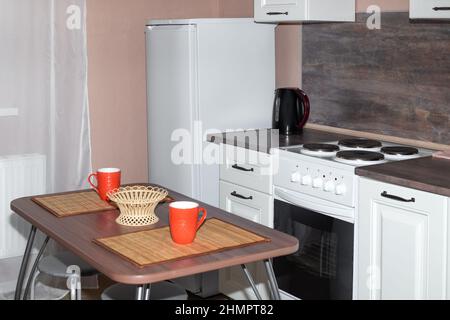 Das Innere einer kleinen gemütlichen Küche in der Wohnung. Holztisch, Kühlschrank und Küche in weiß. Stockfoto