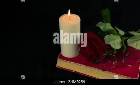 Eine rote Rose mit einem Buch und einer brennenden Kerze auf schwarzem Hintergrund. Von oben brennende Kerze mit frischem Rot mit einer Rose auf dem Buch auf schwarzem Hintergrund Stockfoto
