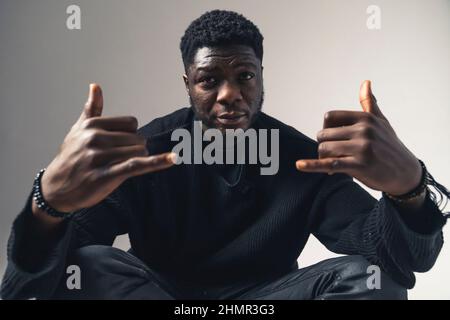 Schauriger Afroamerikaner sitzt mit shaka-Schild, das auf den weißen Hintergrund der Kamera starrt - Porträtaufnahme. Hochwertige Fotos Stockfoto