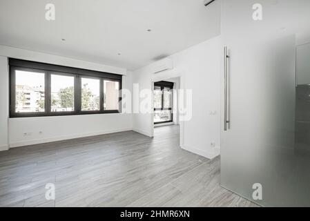 Leeres Wohnzimmer in einem Loft mit großen schwarzen Aluminiumfenstern mit grauen holzähnlichen Keramikböden und einer lichtundurchlässigen Glasschiebetür Stockfoto