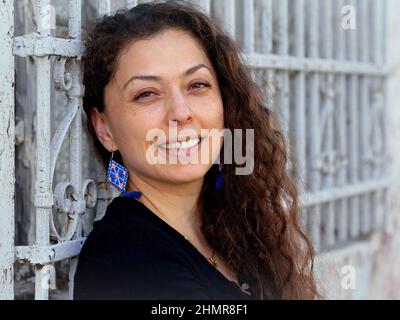 Schöne positive junge mexikanische kaukasische Frau mit langen Locken lächelt vor einem weißen schmiedeeisernen Fenstergrill und schaut auf den Betrachter. Stockfoto