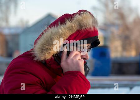 Ein junger Mann in einer roten Daunenjacke mit Brille spricht auf einem Handy Stockfoto