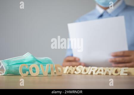 Broker präsentiert die Covid-Versicherung. Holztext mit grüner medizinischer Maske. Objekt befindet sich auf dem Tisch mit Platz an der Wand. Diese Aufnahme war auf der Vorderseite fokussiert Stockfoto