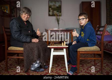 Alte Dame, die mit ihrem Enkel im Schach zu Hause spielt, im Lebensstil. Großmutter und kleiner Junge zeigen Daumen hoch, um Schach zu spielen. Stockfoto