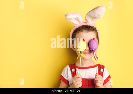 Portrait of funny Little girl covers Eyes with two Easter Eggs, wears ohres, posiert isoliert über gelbem Studio-Hintergrund mit Kopieplatz für Anzeige Stockfoto