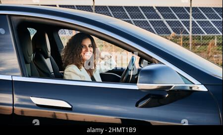 Attraktive junge Frau, die die Kamera anschaut und lächelt, während sie ein Elektrofahrzeug fährt. Schöne Geschäftsfrau mit lockigen Haaren sitzt auf dem Fahrersitz im Elektroauto. Stockfoto