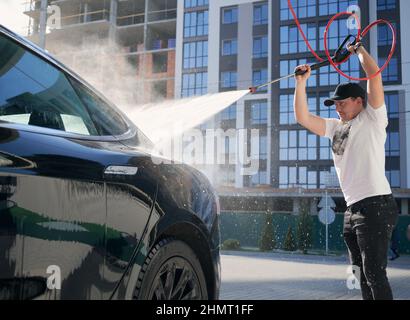 Mann mit Schutzkappe, der sein schwarzes modernes Auto mit Hochdruckwasser in Betrieb genommen hat. Waschen von Auto mit fließendem Wasser auf dem Hintergrund des Wohngebäudes. Stockfoto