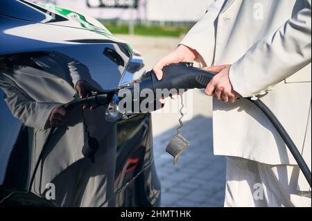 Nahaufnahme der Hände einer Frau, die den Stromversorger an ein Elektroauto anschließt. Prozessladung ökologischer moderner Elektrofahrzeuge. Alternative Energie für den Transport. Stockfoto