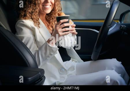 Das verkürzte Porträt einer fröhlichen Geschäftsfrau, die während der Zeit für das Fahren eines Firmenautomobils frühstückt, einer fröhlichen Unternehmerin mit Kaffee zum Mitnehmen und einem Croissant, der vor der Kamera lächelt Stockfoto