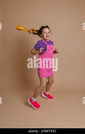 Ausgelassene, aktiv springende kleine Kätzin mit zwei gelben Kanekalon-Zöpfen, die mit einem Lächeln mit fehlenden Zähnen in rosa Jumpsuit, lila, auf die Kamera blicken Stockfoto