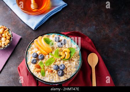 Haferbrei mit Joghurt, Beeren und Nüssen auf burgunderfarbener Serviette. Schüssel mit Haferflocken, Blaubeeren, Äpfeln und Honig auf dunklem Grund. Gesunde Brea Stockfoto