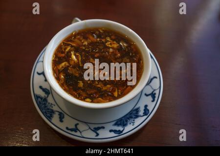 Chinesische würzige und saure Suppe in einer weißen Porzellanschüssel mit asiatischem Muster auf einem dunkelbraunen Tisch, Kopierraum, ausgewählter Fokus, enge Schärfentiefe Stockfoto