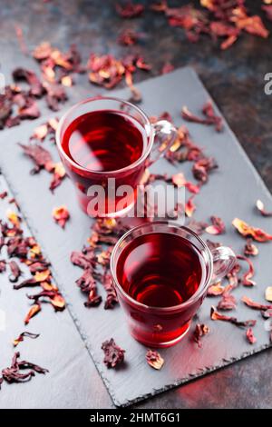 Hibiskus-Tee in Glaskrug auf dunklem Hintergrund. Tasse roten Hibiskustee und trockene Hibiskusblüten auf einer Schiefertafel Stockfoto