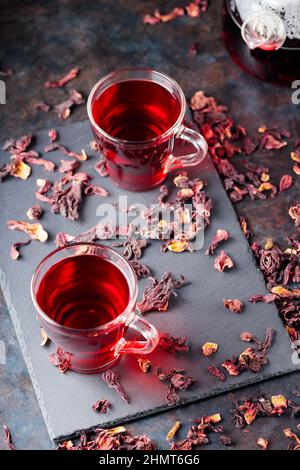 Hibiskus-Tee in Glaskrug auf dunklem Hintergrund. Tasse roten Hibiskustee und trockene Hibiskusblüten auf einer Schiefertafel Stockfoto