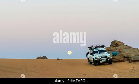 Der alte Toyota Landcruiser parkte an einem Felsberg in der Sahara mit Sand im Vordergrund. Farbenfrohe Morgendämmerung mit einem Vollmond, der im Hintergrund aufgeht. Stockfoto