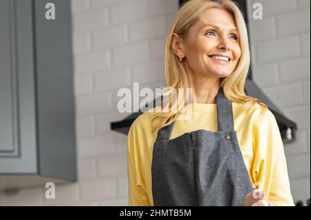 Porträt einer positiven, blonden Frau mittleren Alters in Schürze, die durch das Fenster schaut, Hausfrau, die zu Hause in der Küche steht Stockfoto