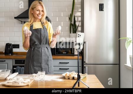 Lebensmittel-Vlogs. Eine Frau mittleren Alters, die Kochvideos in der Küche aufzeichnet, eine Bloggerin siebt das Mehl auf dem Tisch und blickt mit einem Lächeln auf die Kamera, die live per Smartphone streamt Stockfoto