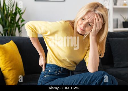 Die blonde Frau fühlte starke Schmerzen im Lendenwirrwarz, der zu Hause auf dem Sofa saß, die besorgte kaukasische Dame hielt eine Hand auf dem unteren Rücken und litt unter dem Gesichtsausdruck Stockfoto