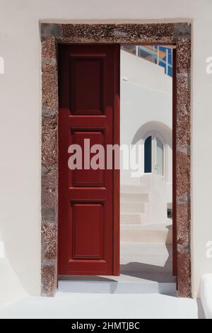 Halb geöffnete wunderschöne rote Tür an der weißen Fassade der griechischen Architektur in Oia, Santorini, Griechenland, Europa. Wunderschöne Details der Insel Stockfoto