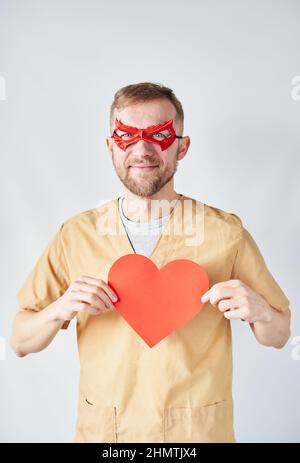 Fröhlich kaukasischen männlichen Kardiologen Arzt in medizinischen Uniform und rote Superhelden-Maske mit rotem Papier Herzform. Erwachsene Arzt Chirurg im Krankenhaus posiert über der Wand. Hochwertige Fotos Stockfoto