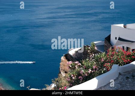 Weiße Architektur mit rosa und roten Blumen auf Santorini, Griechenland. Schöne Landschaft mit Meerblick mit Boot und Wellen. Aussichtsplattform in l Stockfoto