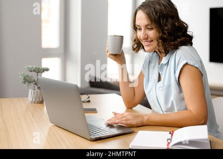 Freiberuflerin träumt bei einer Tasse Kaffee am Morgen, lächelnde Frau sitzt mit einem Laptop am Tisch, schaut auf den Bildschirm und wartet auf Inspiration oder neue Ideen Stockfoto