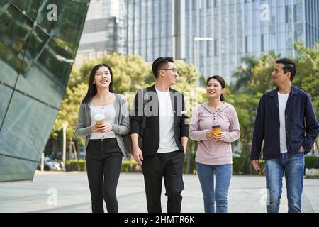 Eine Gruppe von vier jungen asiatischen Menschen, die sich unterhalten und auf der Straße in der modernen Stadt fröhlich und lächelnd spazieren gehen Stockfoto