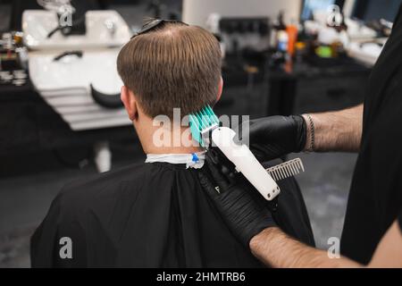 Mann, der sich im Friseurladen Haare schneiden lassen kann. Männlicher Barbier, der mit dem Haarschneider Haare auf den Kopf des Kunden schneidet Stockfoto