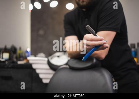 Beschnittenes Bild eines männlichen Barbiers, der seine Rasierklinge in den Händen hält Stockfoto