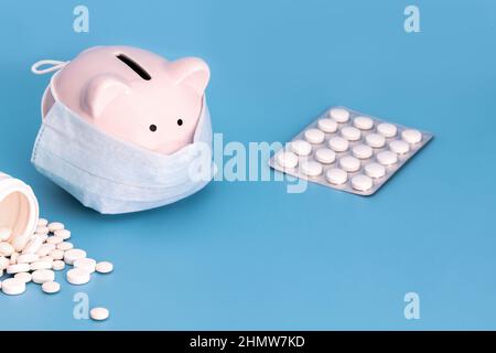 Pink Piggy Bank steht auf einem blauen medizinischen Hintergrund mit einer medizinischen Maske, viele weiße Pillen verstreut herum. Chinesisches Coronavirus, 2019-nCoV Stockfoto