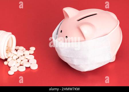 Pink Piggy Bank steht auf einem leuchtend roten Hintergrund und trägt eine medizinische Gesichtsmaske, auf der linken Seite sind weiße Tabletten aus einem weißen Glas verstreut. Geldbox. Chi Stockfoto
