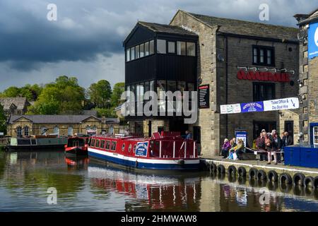 Männer, Frauen, die sich auf einem roten schmalen Boot (Ticketschalter, Anlegestellen am Kanal) für ein touristisches Freizeiterlebnis anstellen - Leeds-Liverpool Canal, Yorkshire, England, Großbritannien. Stockfoto