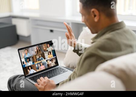 Urkomischer Mann, der ein Notebook für Videoverbindungen verwendet, während er zu Hause bleibt. Lächelnder, multirassischer Kerl winkt Hallo, grüßt Kollegen und nutzt die Computer-App für Videoanrufe Stockfoto