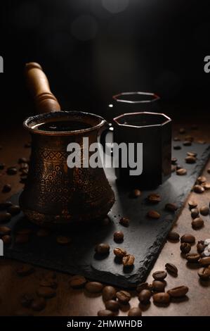 Heißer Kaffee in einem Türken. Der Morgenkaffee wird auf einem schwarzen Brett serviert. Auf der rechten Seite befinden sich zwei Tassen des Getränks. Schwarze Kaffeebohnen sind überall verstreut Stockfoto