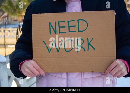 Arbeitslose Frau, die einen Karton mit Inschrift HÄLT, BRAUCHT ARBEIT. Konzept des Arbeitsplatzverlustes und der Suche nach einem neuen. Sie hat finanzielle Probleme. Stockfoto