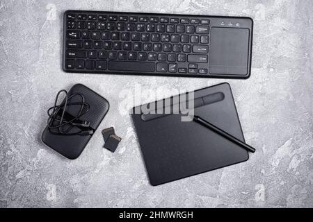 Draufsicht auf Designer-Arbeitsplatz mit Grafiktablett, Tastatur und tragbarer Festplatte auf grauem Beton-Hintergrund Stockfoto