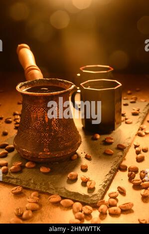 Heißer Kaffee in einem Türken. Der Morgenkaffee wird auf einem schwarzen Brett serviert. Auf der rechten Seite befinden sich zwei Tassen des Getränks. Schwarze Kaffeebohnen sind überall verstreut Stockfoto