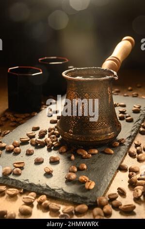 Heißer Kaffee in einem Türken. Der Morgenkaffee wird auf einem schwarzen Brett serviert. Auf der rechten Seite befinden sich zwei Tassen des Getränks. Schwarze Kaffeebohnen sind überall verstreut Stockfoto